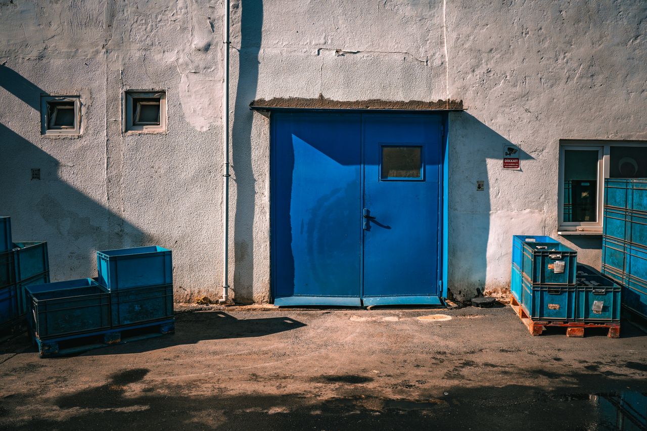 about-07 An industrial blue door surrounded by blue storage crates on a sunny day.